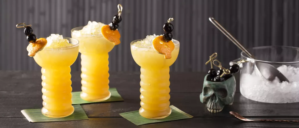 Three Glasses of Zombie garnished with crushed ice, orange peel and cherries on coaster alongside pot of crushed ice with spoon, cherries in skull shaped pot and a bar spoon