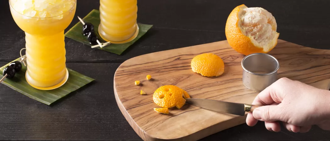a person cutting orange peel with knife into skull shape on tray alongside an orange and a circle shape cutter