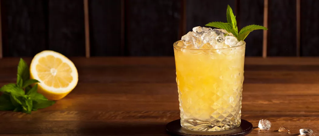 A Double Oak Whiskey Smash in glass on coaster with crushed ice and mint,  alongside lemon half and mint leaves