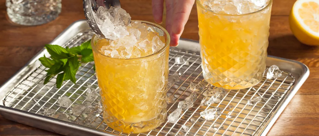 Two glasses on a steel tray: one filled with crushed ice and the other being filled, along with mint leaves