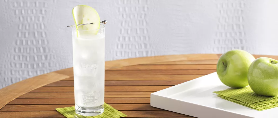 . EFFEN® Green Apple Cooler glass with an apple slice on a green coaster, alongside apples and coasters on a white tray.