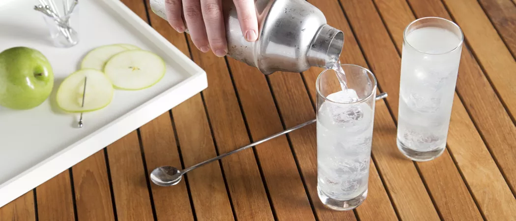 A person pours EFFEN® Green Apple Cooler into a glass filled with ice next to another glass of the same drink, a bar spoon, and a white tray with a green apple, apple slices, and a pin.