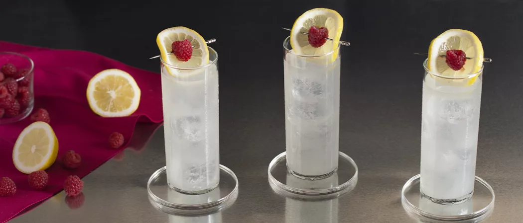 Three glasses of EFFEN® Raspberry Lemonade garnished with lemon slice and raspberry on glass coaster alongside few lemon slices and raspberries on red cloth