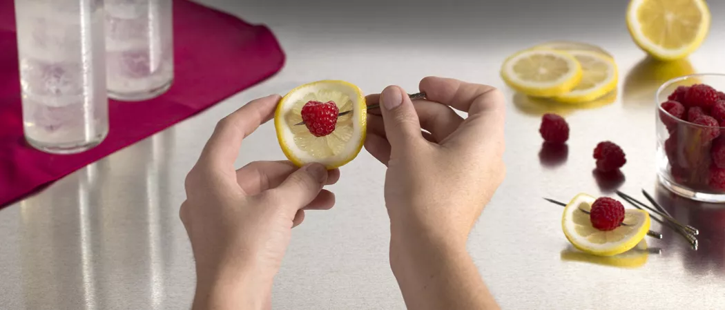 A person creating garnish using lemon slice, respberry and steel toothpick alongside some lemon slices and bowl of raspberries 