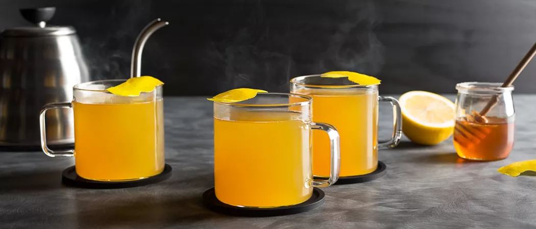 Three cups of Jim Beam® Black Hot Toddy garnished with lemon peel on coaster alongside honey pot with honey dipper and half lemon