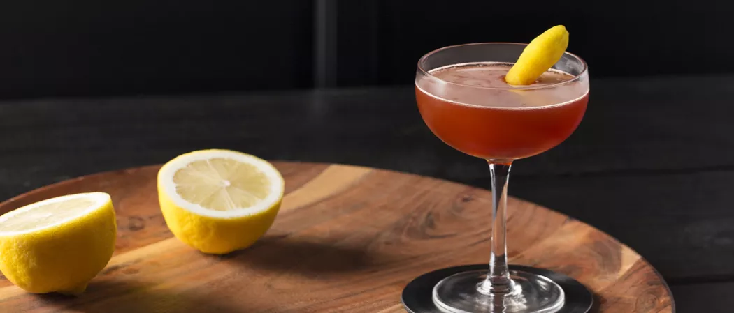 A glass of Black Sunset garnished with lemon peel on coaster alongside two lemons on a round wooden tray