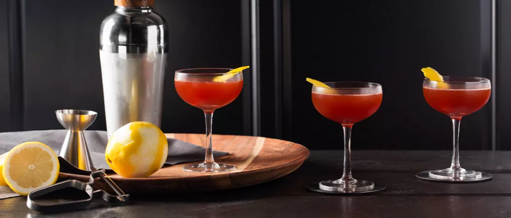 Three glasses of Black Sunset garnished with lemon peel. Two glasses are on the table, and one glass, along with a lemon, jigger, and bottle on a wooden tray.