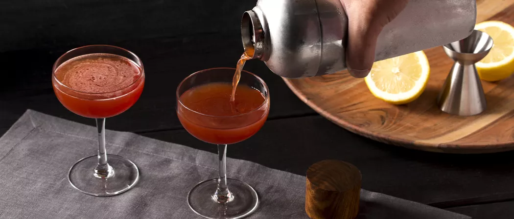 Two glasses of Black Sunset on a gray napkin. One glass has a person pouring the drink from a bottle, alongside  two half lemons and a jigger on a round wooden tray.