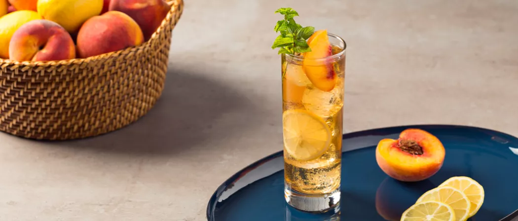 A highball glass of Peach Iced Tea, garnished with lemon wheel, peach slices, and mint leaves, accompanied by halved peach and lemon slices, all on a tray. Nearby, a bucket filled with peaches and lemons.