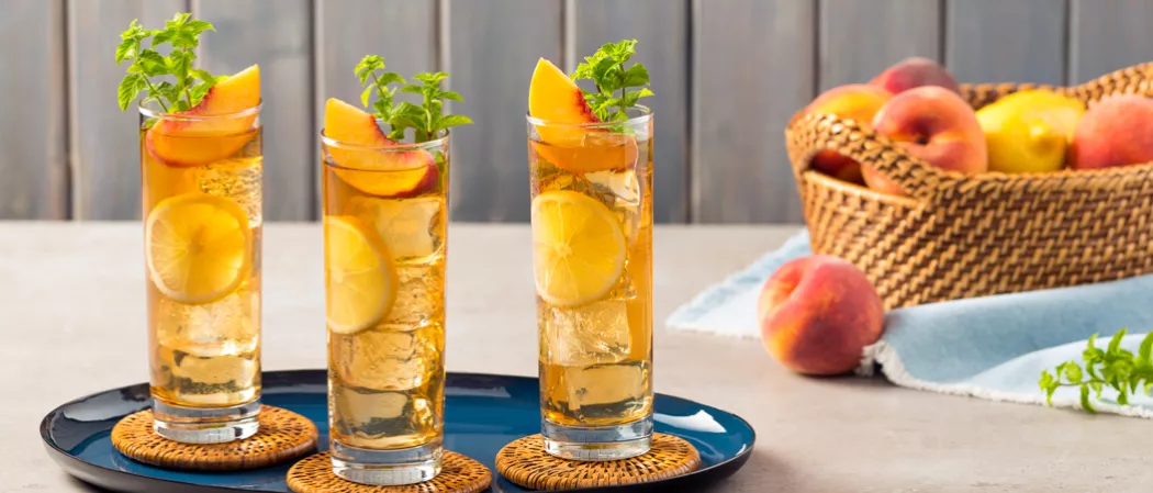 Three highball glasses of Peach Iced Tea, garnished with lemon wheel, peach slices, and mint leaves on a coaster in a tray. A bucket of peaches and lemons on a napkin nearby.