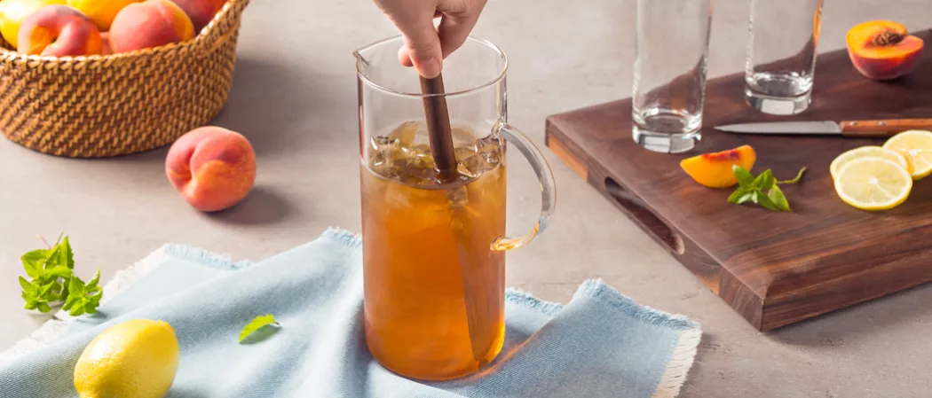 A person stirs Peach Iced Tea in a jug placed on a napkin. Nearby, a bucket of peaches and lemons, a chopping board with 2 empty glasses, a knife, peach pieces, lemon slices, and mint leaves.