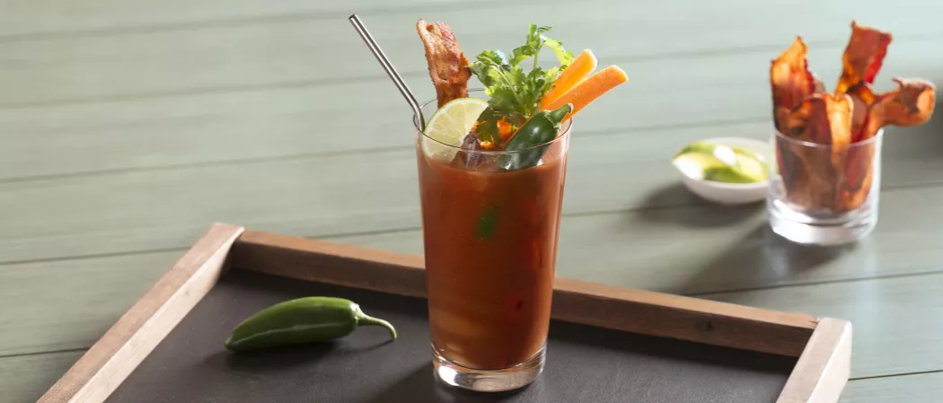 Glass of Sunday Funday Bloody Mary garnished with lime wedge, cilantro, becon slices on wooden tray alongside glass with becon slices