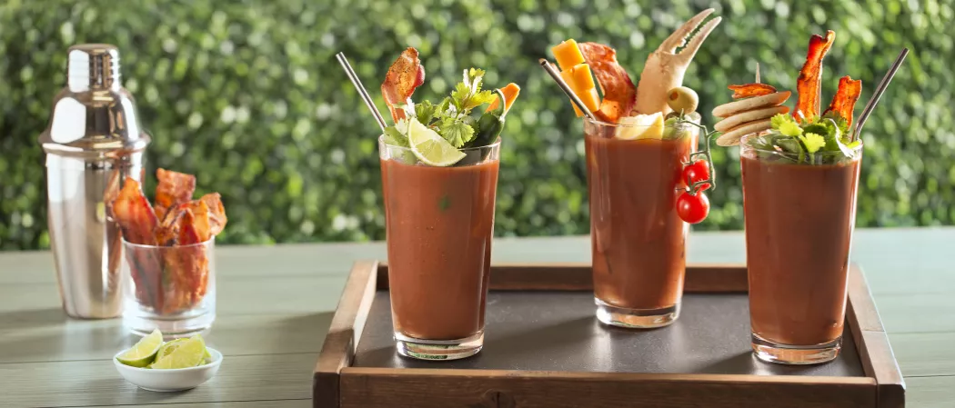 Three glasses of Sunday Funday Bloody Mary on wooden tray garnished with lime wedge, cilantro, becon slices