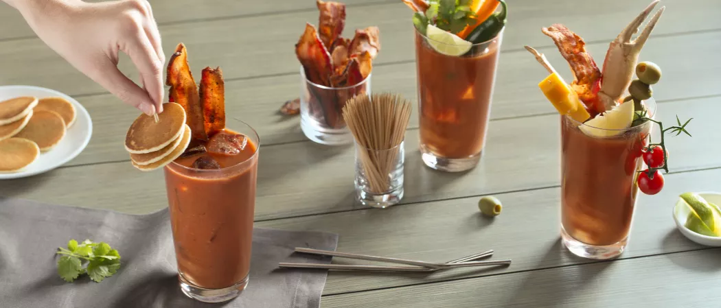 Person garnishing glass of Sunday Funday Bloody Mary with small pancakes alongside two more glasses  of same