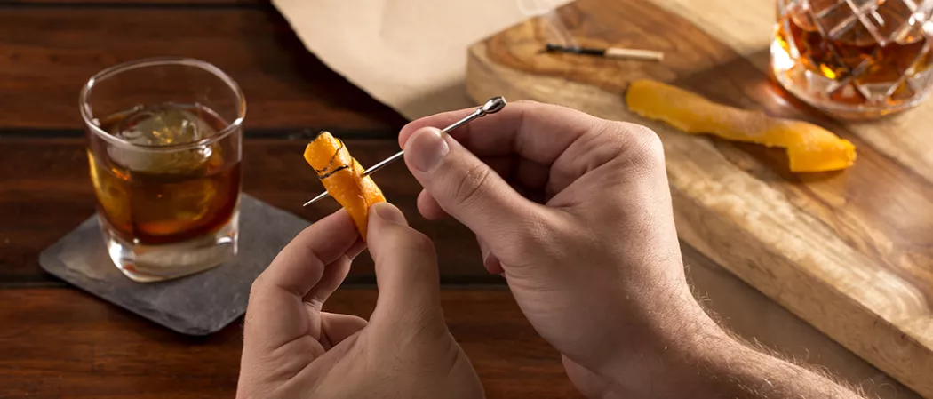 a person putting steel toothpick into orange peel and glass of Rye Opener on coaster in background