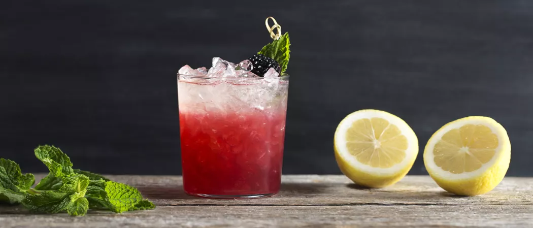 Glass of Independence Spice garnished with skewered mint leaf and blackberry alongside few mint leaves and two half lemons