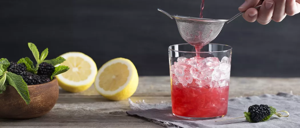 A person Pouring Independence Spice into glass filled with ice alongside bowl of mint leaves and mulberries, two half lemon and a mulberry