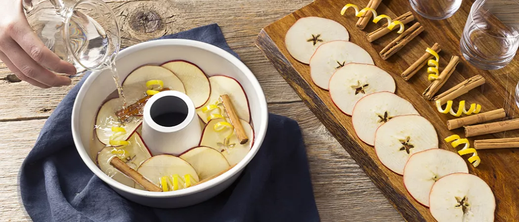 A person pouring water in bowl filled with apple slices, lemon twist and cinnamom sticks alongside wooden board decorated with apple slices, cinnamon sticks and lemon twist