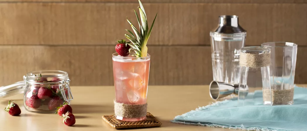 A glass of Bay Breeze with ice cubes, garnished with a pineapple slice and strawsberry on a coaster. Alongside, a bowl of strawberries, Two glasses on a blue cloth with a jigger and a shaker.