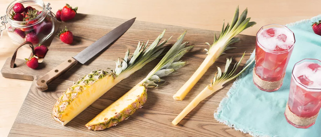A knife and a chopped pineapple with two glasses of Bay Breeze with ice cubes on a blue napkin, all arranged on a chopping board alongside a jar full of strawberries.