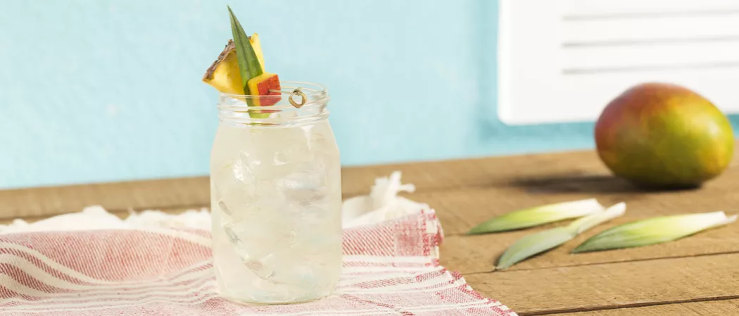 Cruzan® Confusion served in a glass with pineapple, pineapple leaves, and mango on a table with a cloth, next to another mango