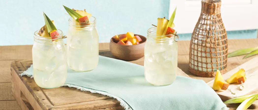 Three glasses of Cruzan® Confusion with pineapple, leaves, mango on a table with a cloth, next .to a bowl of chopped mango, and a glass jar in wooden architecture with 2 garnishing elements ready for use.