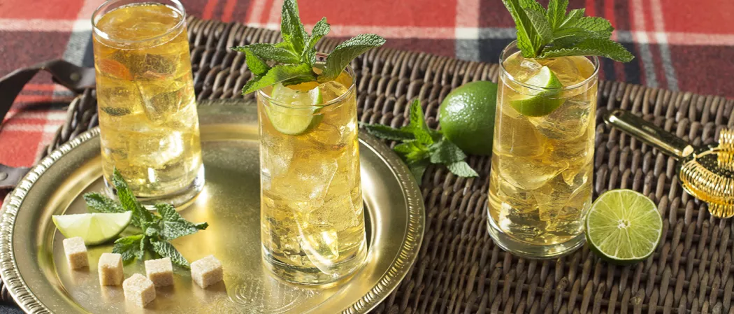 Glass of Derby Spritzer garnished with mint leaves and lime wedge alongside another glass of Derby Spritzer, lime wedge, mint leaves and suger cubes on golden plate alongside glass of Derby Spritzer garnished with mint leaves and lime wedge