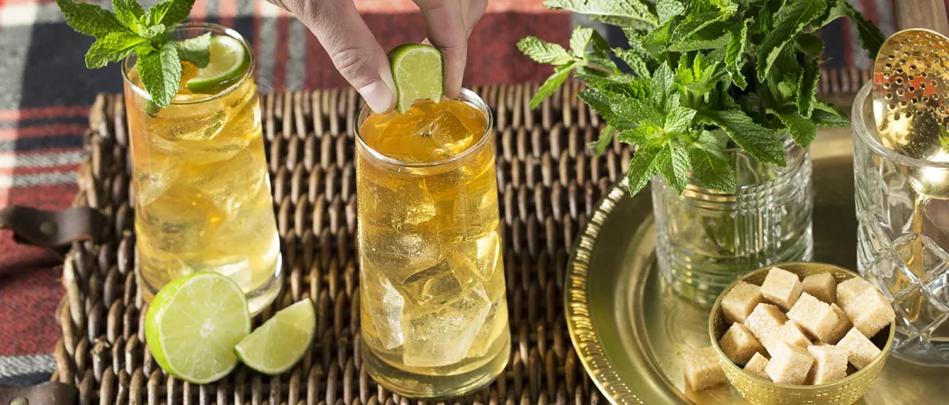 A person squeezing lime into glass of  Derby Spritzer alongside glass of Derby Spritzer garnished with mint leaves and lime wedge, lime and a golden plate with bowl of sugar cubes and pot of mint leaves