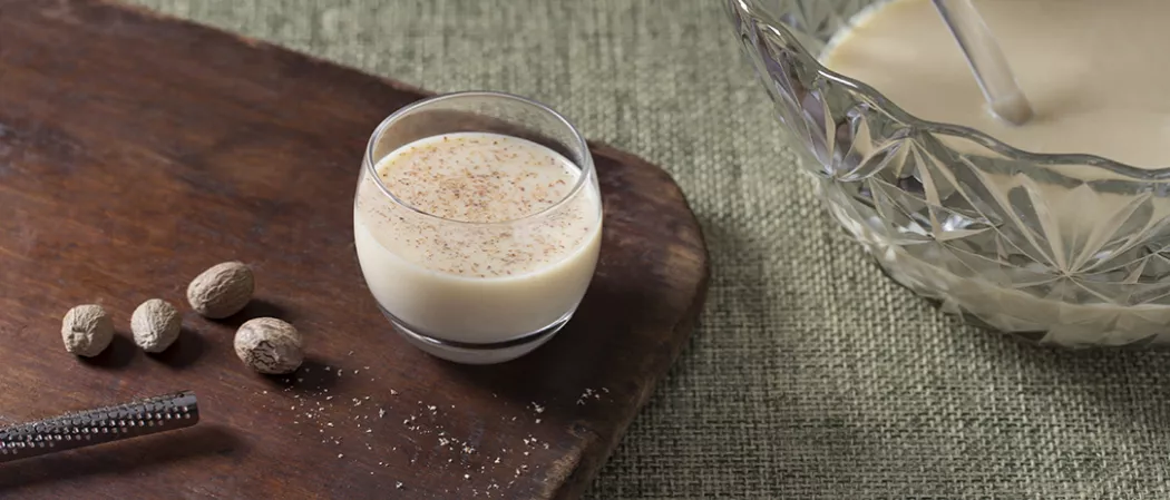 Glass of Yule Nog alongside few nutmegs on wooden table.