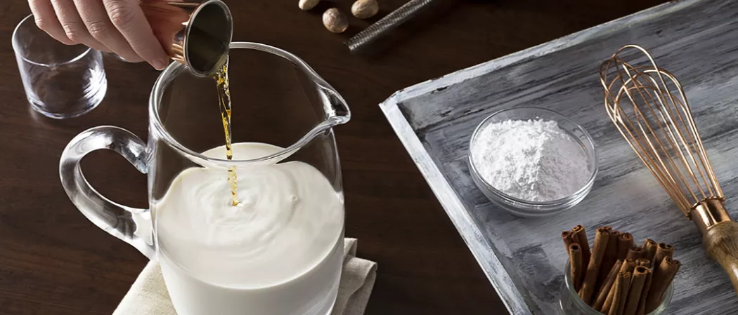 A person pouring Jim Beam® Original into a jar on a napkin, half-filled with milk alongside an empty glass. Next to it is sugar powder, cinnamon sticks, and a balloon whisk on a wooden tray.