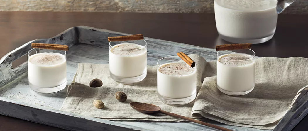 Four glasses of Bourbon Milk Punch garnished with cinnamon sticks. Three glasses placed on napkins with nutmeg, accompanied by a wooden spoon in a wooden tray.