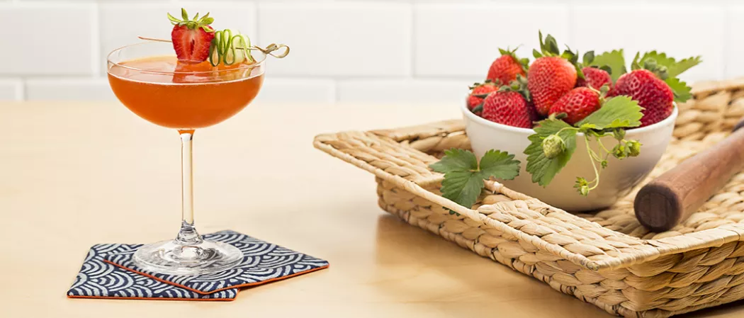 Glass of Melon Berry Mist garnished with strawberry and cucumber wheel on coaster alongside tray with bowl of strawberries
