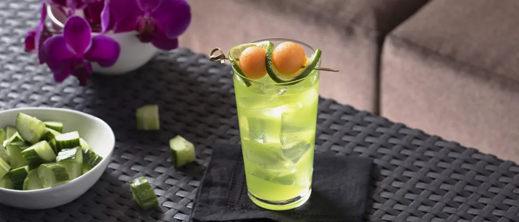 Glass of Melon Cooler garnished with melon ball and lemon slice alongside bowl of diced cucumber