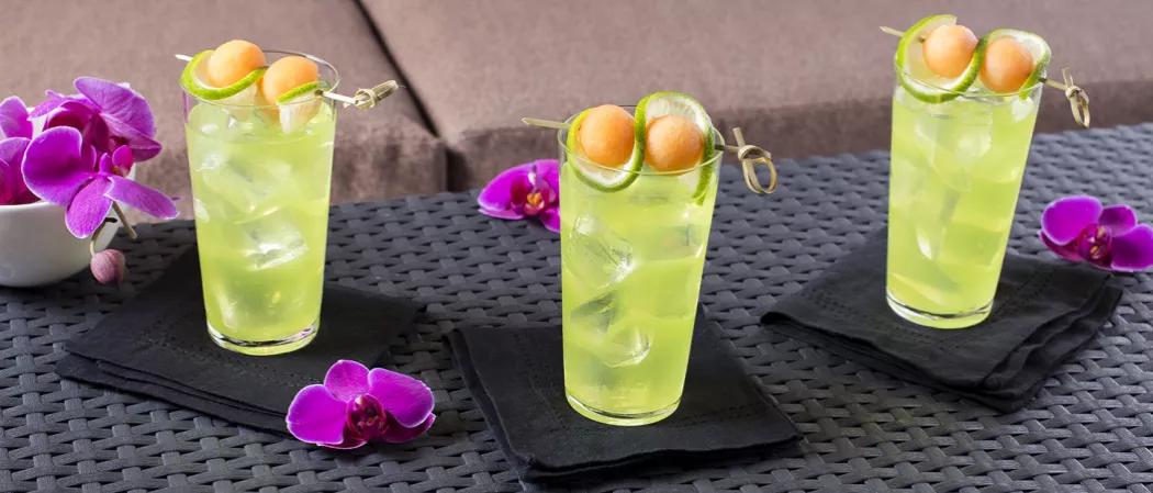 Three glasses of Melon Cooler garnished with melon ball and lemon slice on black coaster