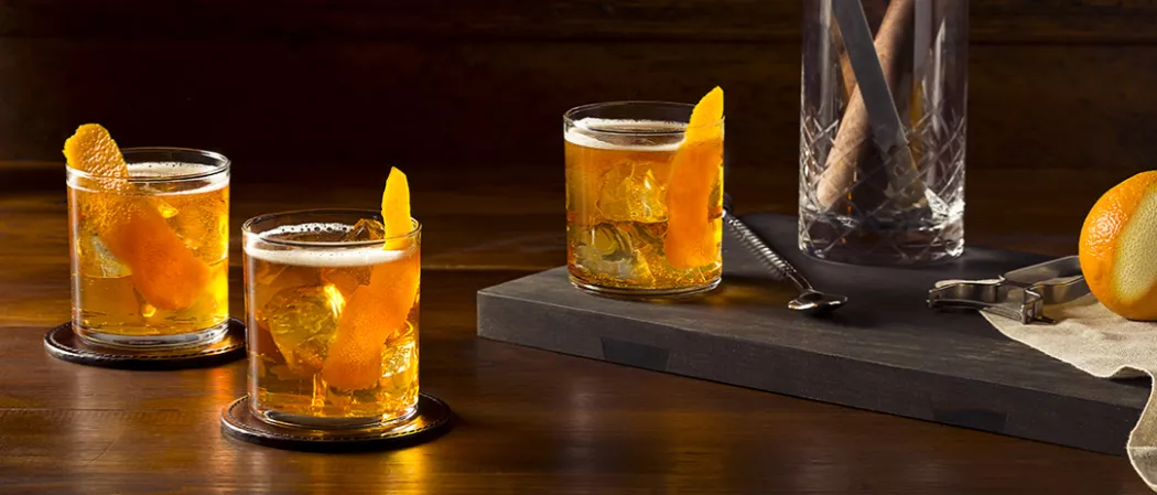 Two glasses of Rye Squeeze garnished with orange peel on coaster alongside glass of Rye Squeeze garnished with orange peel and a bar spoon on board