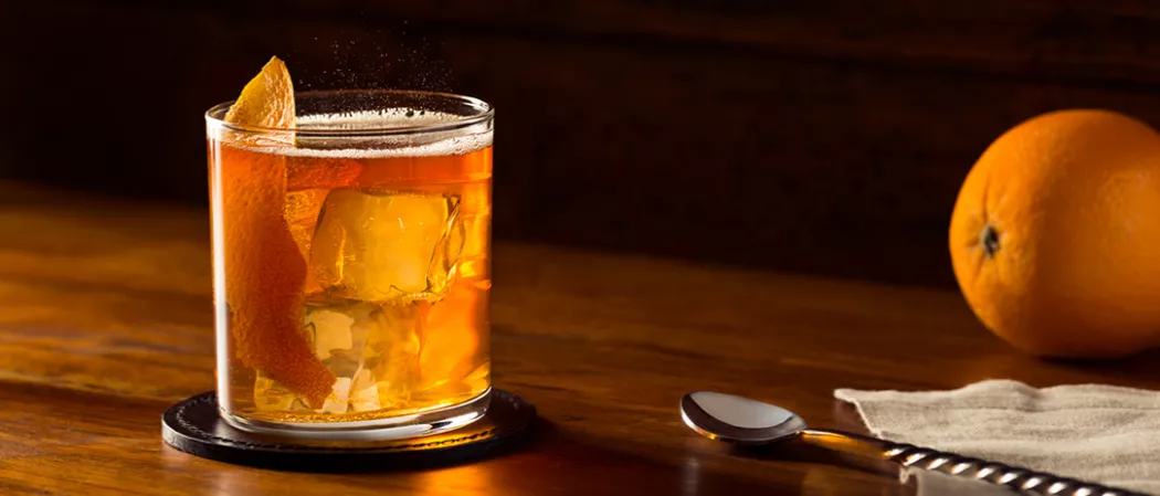 Glass of Rye Squeeze garnished with orange peel on coaster alongside a bar spoon and a orange