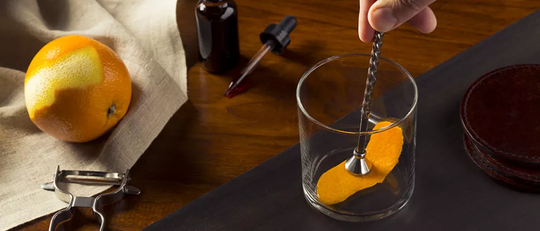 Person muddling orange peel into glass alongside a orange and a peeler