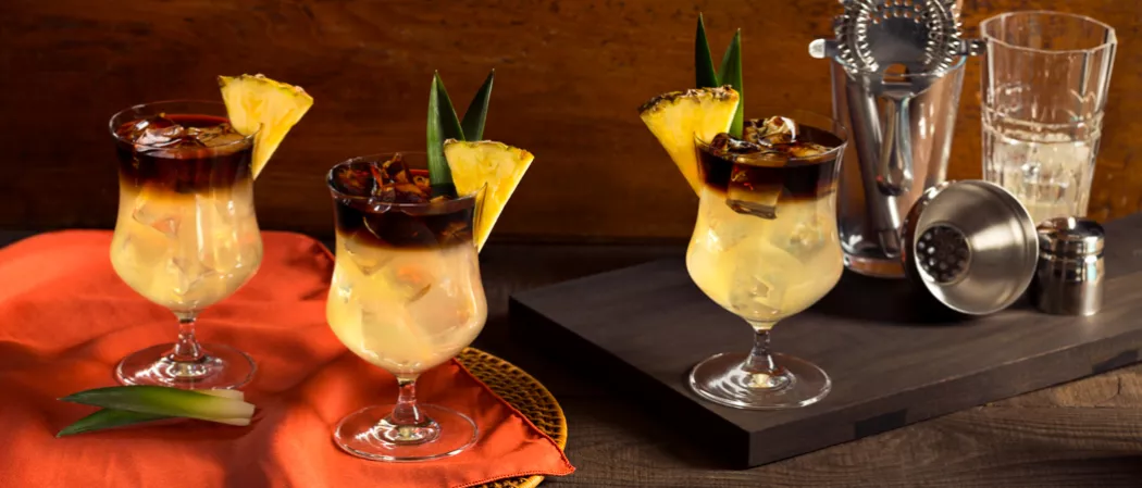 Three Glasses of Cruzan® Mai Tai garnished with pineapple wedge and leaf one on wooden board alongside shaker and strainer in glass