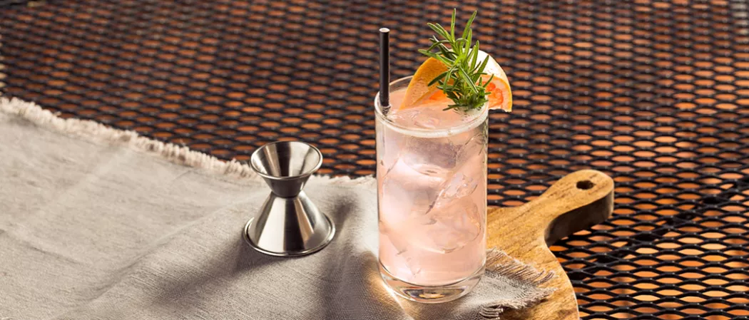 Glass of Paloma garnished with grapefruit wadge, rosemary sprig and straw alongside a jigger
