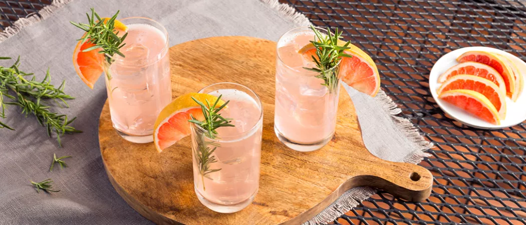 Three glasses of Paloma garnished with rosemary sprig and grapefruit wedge on wooden tray alongside plate of grapefruit wedges