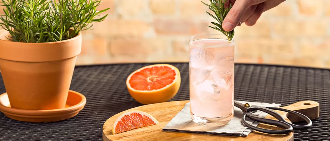 Person garnishing glass of Paloma with rosemary sprig on coaster alongside grapefruit wedge and scissor on wooden tray alongside half grapefruit and rosemary plant pot