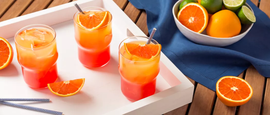 Three Tequila sunrise drinks with orange wedges, two glasses with straws, and a bowl of orange and lemon wedges on a blue cloth