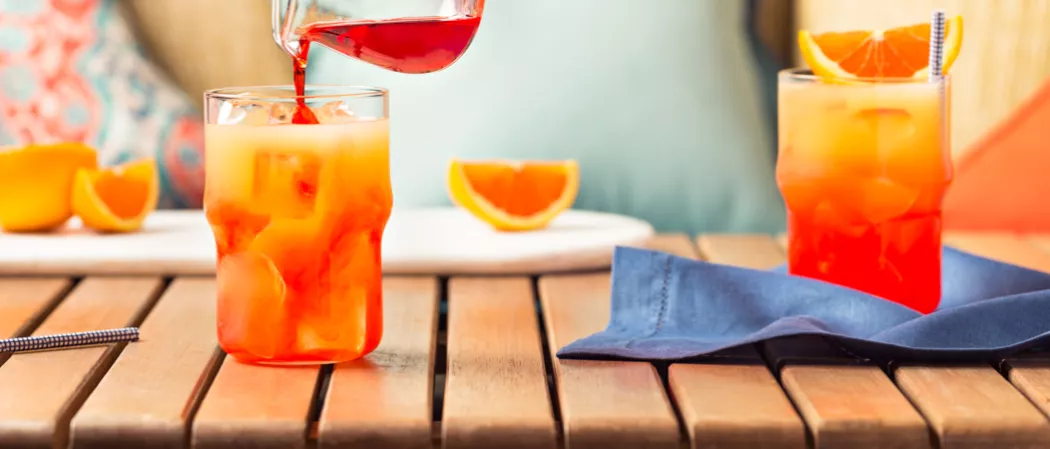 A person pouring Tequila Sunrise from a jar in to a glass, alongside a straw, a glass of Tequila Sunrise, garnished with ice cubes and orange wedges