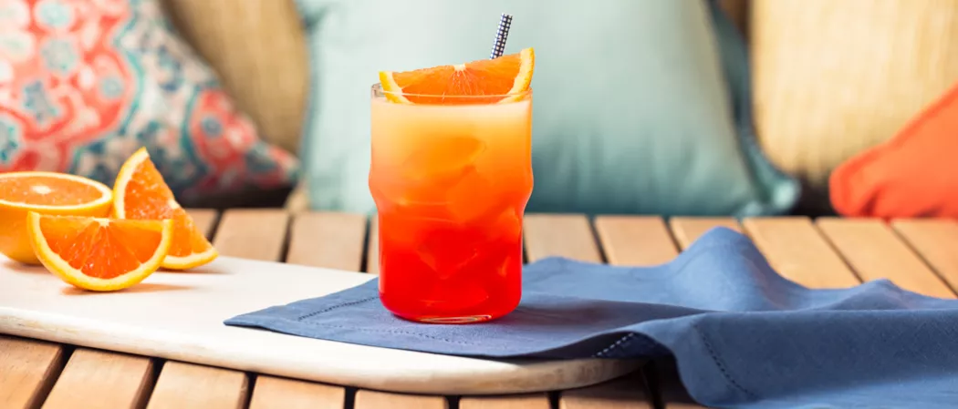 A Glass of Tequila Sunrise, placed on a blue cloth and garnished with lemon and orange wedges, with a straw in it, sits alongside wedges of orange on a white tray