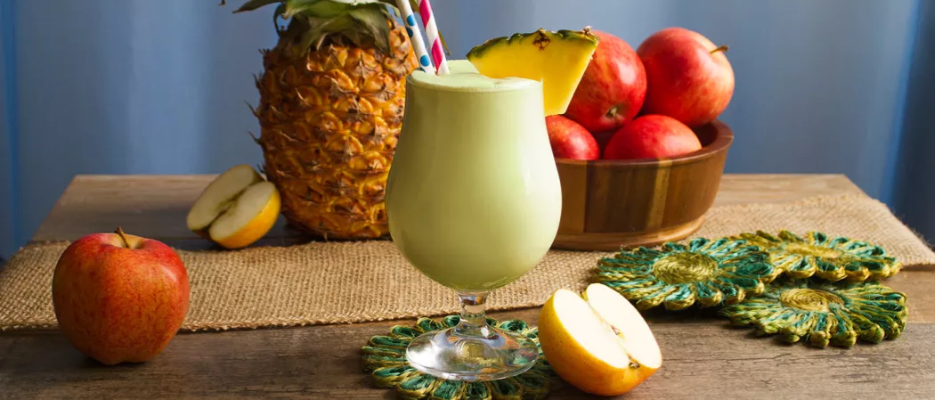 Glass of Apple Daiquiri garnished with pineapple slice and strawss on a coaster alongside few coasters, apples, a pineapple and a basket of apples