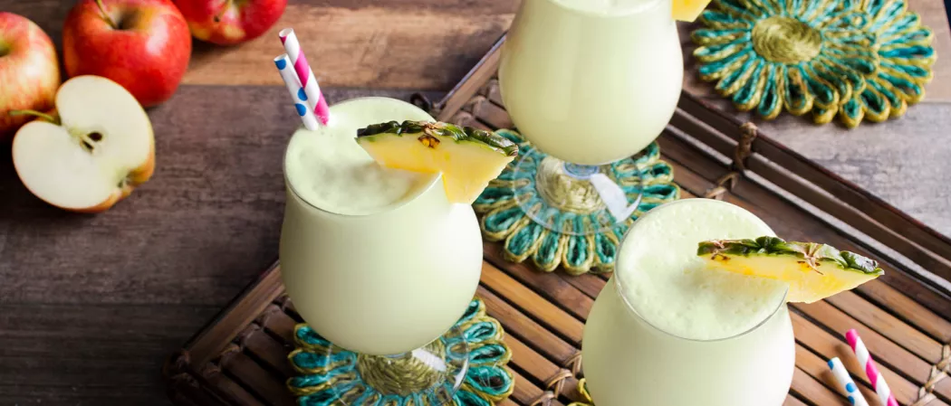 Three glasses of Apple Daiquiri garnished with pineapple slice and strawss on a coaster on wooden tray alongside few apples