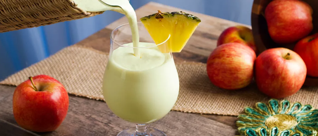 pouring  Apple Daiquiri into a glass garnished with pineapple slice alongside a coaster and some apples.