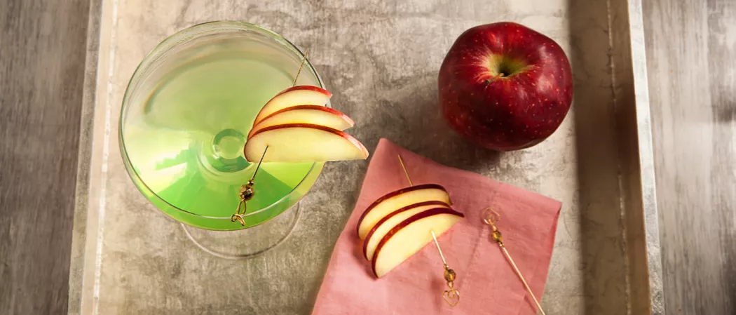 A Glass of Appletini, each garnished with an apple slice pierced on a cocktail pick on the rim, are accompanie by slices of apple pierced with cocktail picks, and few loose cocktail picks, all on a red cloth within a wooden tray.