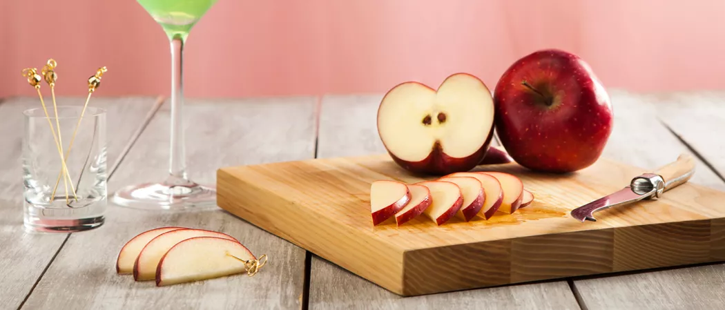 An apple and a few slices of apple, accompanied by a knife, are placed on a wooden table. Additionally, there are a few slices of apple pierced with cocktail picks and a few loose cocktail picks in a glass
