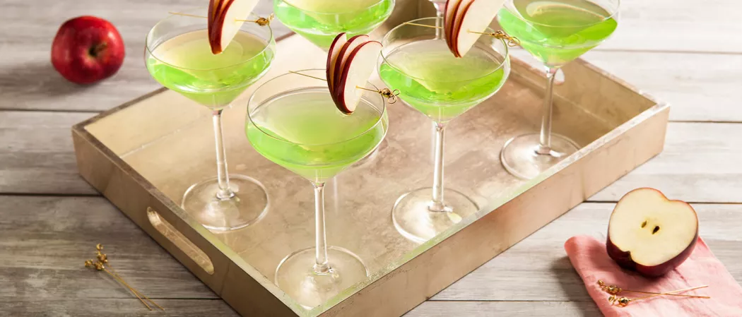Five glasses of Appletini, each garnished with an apple slice pierced on a cocktail pick on the rim of the glass, are arranged in a wooden tray. Additionally, slices of apple and cocktail picks on a red cloth.